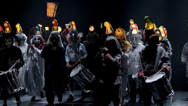 Videostandbild aus dem Video "Morgestraich, 2022" von Clément Cogitore: Darsteller in Kostüm mit Trommeln auf einer dunklen Bühne