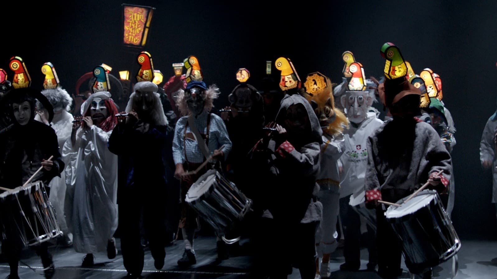 Videostandbild aus dem Video "Morgestraich, 2022" von Clément Cogitore: Darsteller in Kostüm mit Trommeln auf einer dunklen Bühne