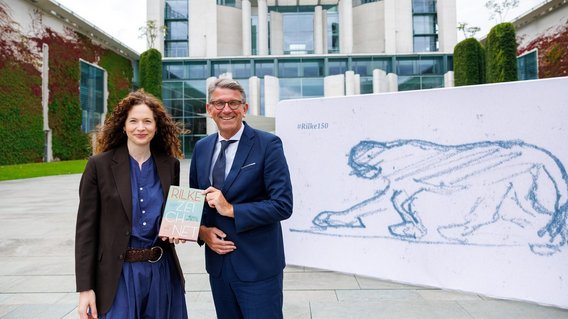 Staatsminister Wolfram Weimer mit Sandra Richter vor einer Stellwand mit der Zeichnung Rilkes und dem Bundeskanzleramt