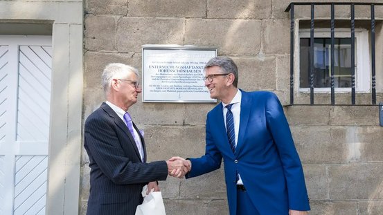 Kulturstaatsminister Weimer und Helge Heidemeyer, Direktor der Gedenkstätte Berlin-Hohenschönhausen reichen sich die Hand, im Hintergrund ist die enthüllte Gedenktafel zu sehen. 