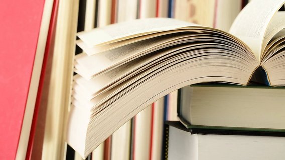 Ein aufgeschlagenes Buch auf einem B&uuml;cherstapel, im Hintergrund weitere B&uuml;cher. 