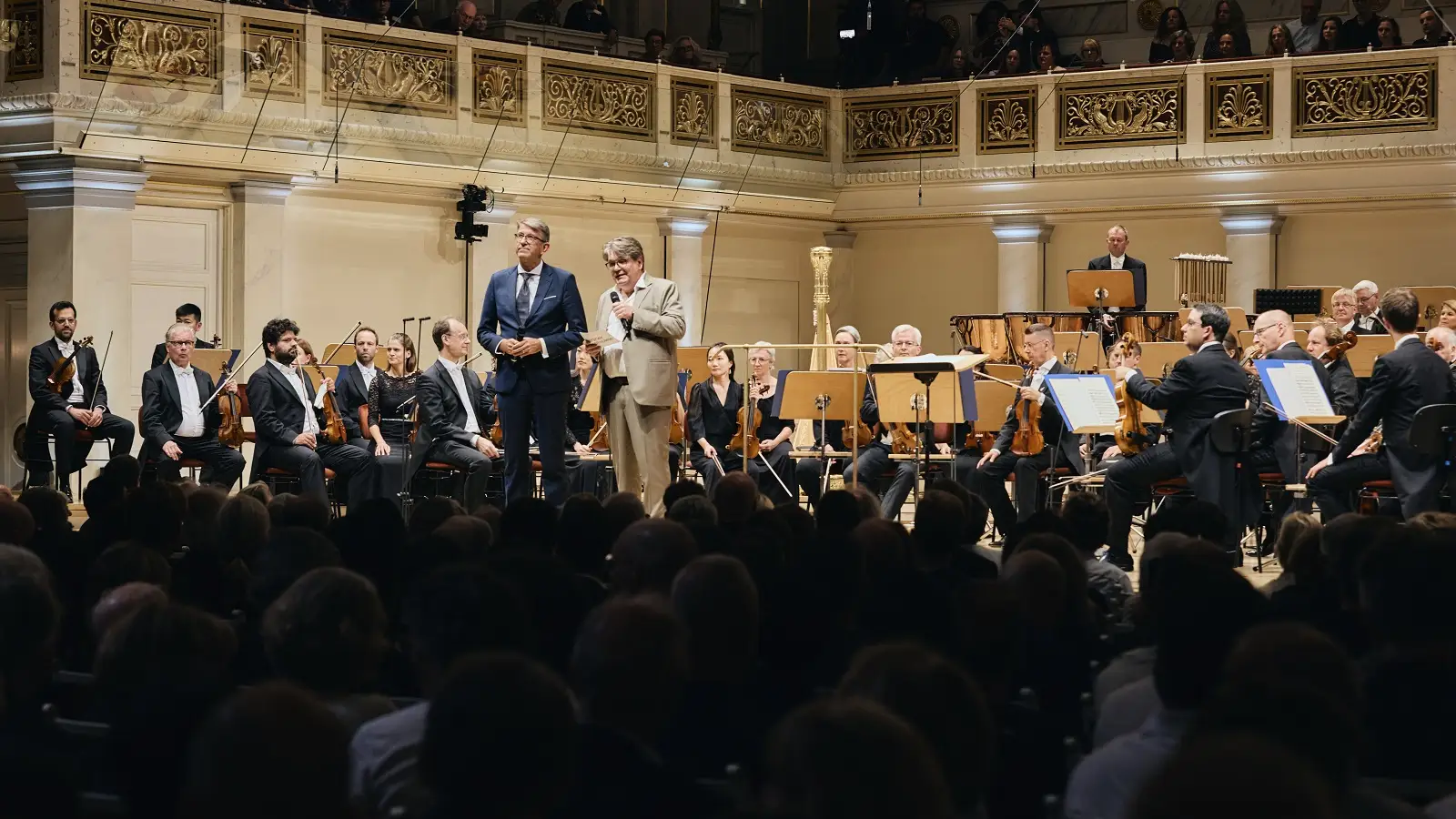 Staatsminister Wolfram Weimer steht auf der hell beleuchteten B&uuml;hne, im Hintergrund sitzt das Orchester. 